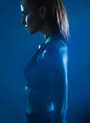 Starke junge Frau in Sportkleidung vor blauem Hintergrund. Selbstbewusste junge Sportlerin aus der Seitenansicht. - JLPSF14572