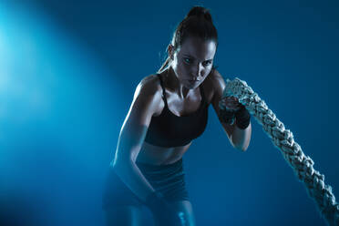 Sportler bewegt die Seile in Wellenbewegung als Teil des Fettverbrennungstrainings. Sportlerin trainiert mit Kampfseilen auf blauem Hintergrund. - JLPSF14586