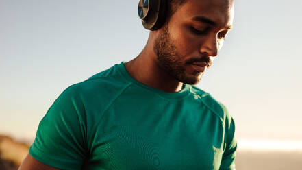 Close up of an athlete standing outdoors wearing headphones. Fitness man listening to music while running. - JLPSF14599