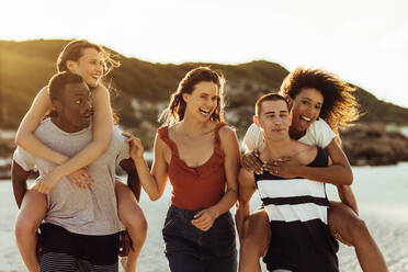 Cheerful friends running a piggy riding race on a beach. Multiethnic friends enjoying and having fun on holiday. - JLPSF14640