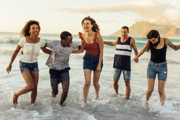 Friends on vacation playing in the sea. Multiethnic men and women friends running in the sea water holding each other. - JLPSF14641