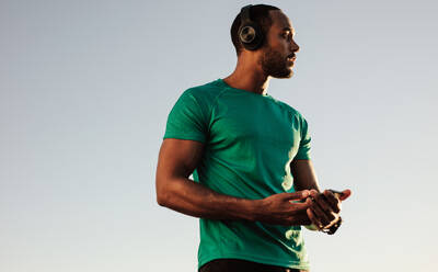 Close up of an afro american man in tshirt standing outdoors. Male athlete listening to music during workout. - JLPSF14649