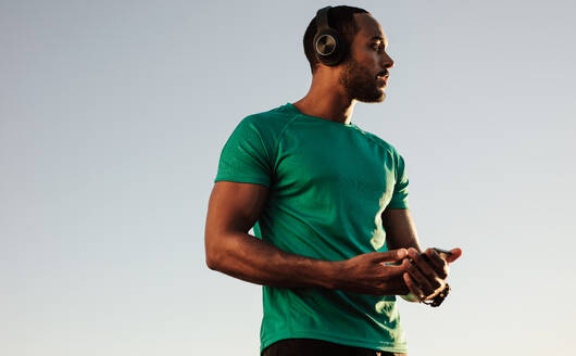 Nahaufnahme eines afroamerikanischen Mannes im T-Shirt, der im Freien steht. Männlicher Sportler, der beim Training Musik hört. - JLPSF14649