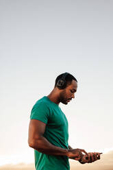 Portrait of a male athlete listening to music on headphones during workout. Muscular fit man using mobile phone standing outdoors. - JLPSF14650