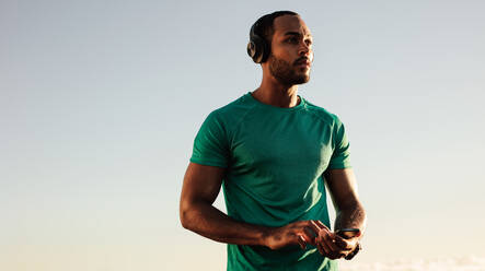 Close up of an afro american man in fitness wear standing outdoors holding a mobile phone. Male athlete listening to music using wireless headphones. - JLPSF14651