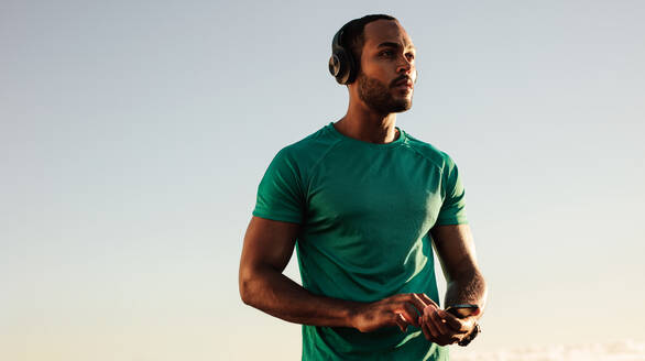 Nahaufnahme eines afroamerikanischen Mannes in Fitnesskleidung, der im Freien steht und ein Mobiltelefon in der Hand hält. Männlicher Sportler, der mit drahtlosen Kopfhörern Musik hört. - JLPSF14651