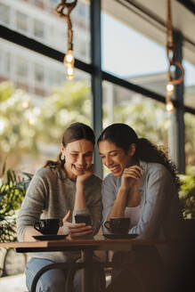 Freunde sitzen in einem Café, schauen auf ein Smartphone und lächeln. Zwei Frauen treffen sich in einem Kaffee und benutzen ein Mobiltelefon. - JLPSF14666