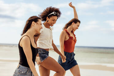 Side view of three young women walking on beach. Cheerful friends in playful mood walking together on the beach. - JLPSF14683