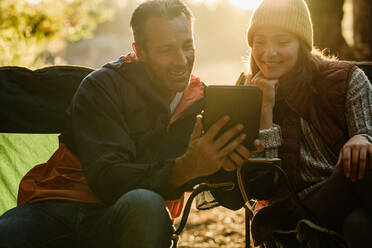 Lächelnder Mann und lächelnde Frau, die sich neben dem Zelt ausruhen und ein digitales Tablet benutzen. Camper mit digitalem Tablet. - JLPSF14693