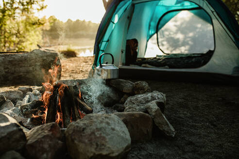 Campsite with a tent, kettle and burning fire pit in countryside. Camping by a lake in forest. - JLPSF14696