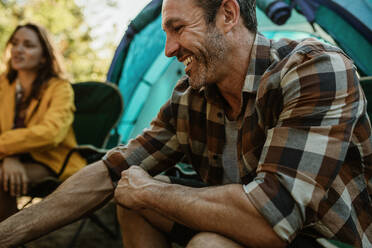 Älterer Mann sitzt auf dem Campingplatz mit einer Frau im Hintergrund. Paar auf dem Campingplatz sitzt außerhalb des Zeltes. - JLPSF14697