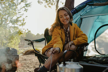 Smiling woman sitting at campsite. Female in warm wear sitting buy campfire outside the tent. - JLPSF14698