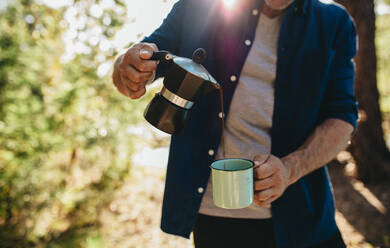 Ausschnitt eines Mannes, der Kaffee aus einem Wasserkocher in eine Tasse gießt. Mann beim Kaffeetrinken auf dem Campingplatz - JLPSF14702
