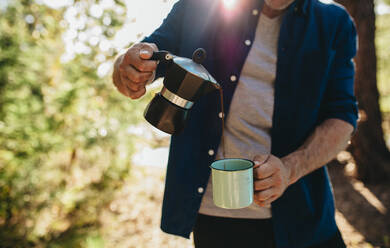 Cropped shot of a man pouring coffee from kettle into a cup. Man having coffee at camping site - JLPSF14702