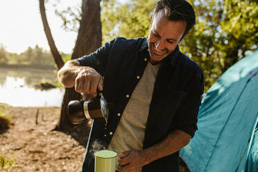 Lächelnder reifer Mann, der Kaffee aus dem Kessel in eine Tasse gießt. Mittlerer erwachsener Mann mit frischem Kaffee auf einem Campingplatz am See. - JLPSF14703