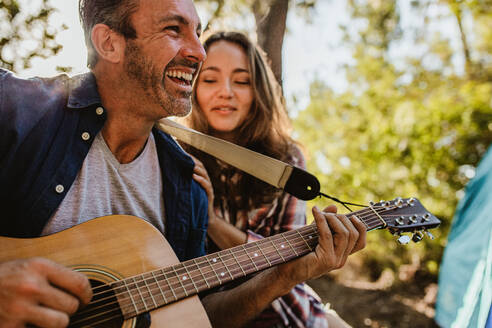 Lächelnder reifer Mann spielt Gitarre mit einer daneben sitzenden Frau. Glückliches reifes Paar beim Camping in der Natur. - JLPSF14707