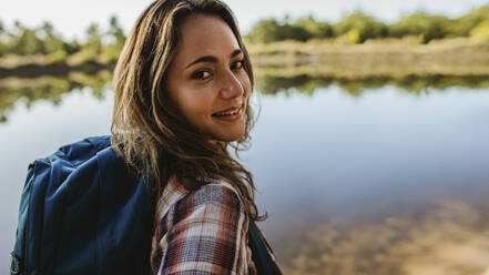Nahaufnahme einer schönen Frau mit Rucksack, die am See steht. Weiblicher Camper schaut in die Kamera. - JLPSF14708
