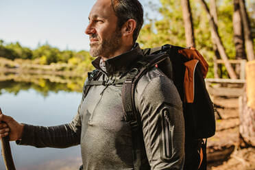 Mittlerer erwachsener Mann mit Rucksack, der am See steht und einen Stock hält. Männlicher Camper, der am See im Wald zeltet. - JLPSF14710