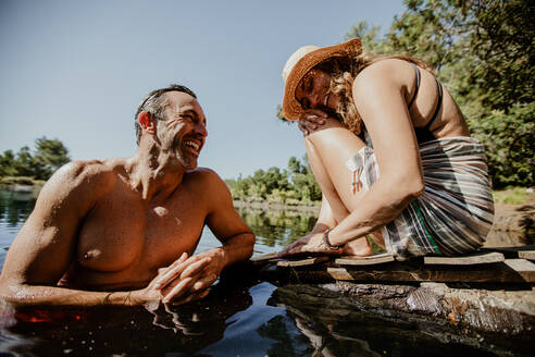 Mature man in lake with woman sitting at the pier. Couple enjoying on their camping vacation at the lake. - JLPSF14721