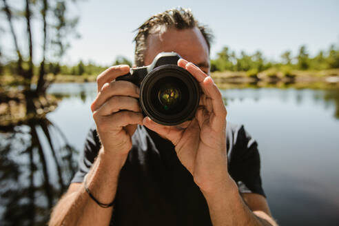 Reifer Mann, der mit einer digitalen Spiegelreflexkamera fotografiert. Männlicher Fotograf, der am See steht und Bilder mit einer Digitalkamera macht. - JLPSF14724