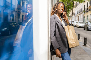 Smiling woman carrying shopping bags talking through mobile phone leaning on wall - DLTSF03290