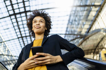 Contemplative businesswoman with smart phone standing on escalator - DIGF19096