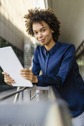 Smiling businesswoman holding tablet PC leaning on railing - DIGF19114