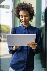 Smiling businesswoman using tablet PC leaning on glass wall - DIGF19117