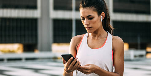 Eine Frau in Sportkleidung benutzt ihr Smartphone nach dem Training. Eine gesunde Frau mit Kopfhörern macht eine Pause vom Training und überprüft ihren Fitnessfortschritt auf dem Handy. - JLPSF14748