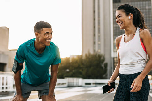 Zwei Menschen machen eine Pause nach dem Training im Freien und lächeln. Mann und Frau entspannen sich nach einem Training und lachen in der Stadt. - JLPSF14749