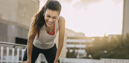 Fitness woman taking a break during the running workout in the city. Fit female athlete smiling and leaning on her knees after morning training session. - JLPSF14753