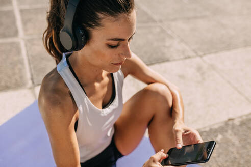 Frau, die beim Sport mit Kopfhörern auf ihrem Smartphone Musik hört. Frau in Sportkleidung, die im Freien sitzt und eine Fitness-App auf ihrem Mobiltelefon nutzt. - JLPSF14772