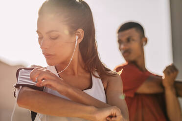 Close-up of a woman listening to music with earphones from her smart phone while exercising in the city with a man in background. Woman using a smartphone to monitor her progress while exercising with her male friend. - JLPSF14785