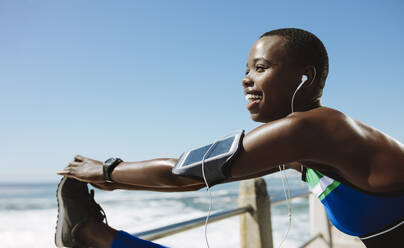 Smiling african woman doing leg stretching exercise on railing at seaside road. Fitness female warming up on the road by the sea. - JLPSF14787