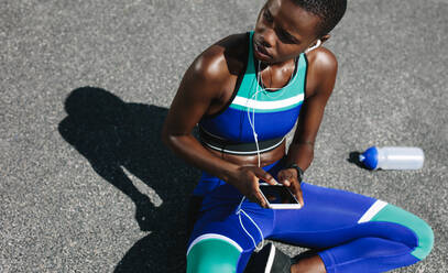 Top view shot of woman with her mobile phone sitting on road after a morning run and looking away. Fit woman on road taking a break after outdoor workout. - JLPSF14790