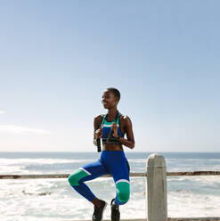 Fröhliche afrikanische Frau in Sportkleidung, die auf einem Geländer an der Küstenstraße sitzt und ein Springseil hält. Frau macht eine Pause nach dem Sport im Freien. - JLPSF14798