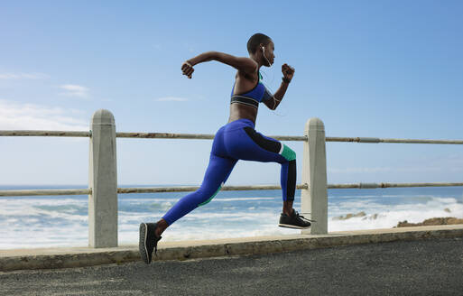 Full length shot of a woman runs on seaside road. Fit female athlete sprinting on seaside promenade in morning. - JLPSF14806
