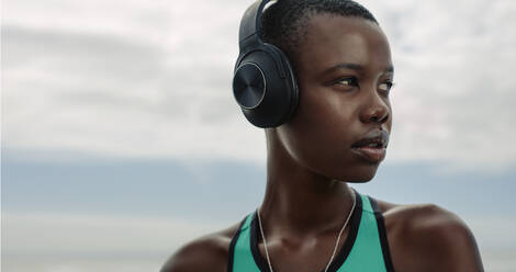 Nahaufnahme einer afrikanischen Sportlerin mit Kopfhörern, die im Freien steht. Frau ruht sich nach dem Training aus und hört Musik über Kopfhörer im Freien. - JLPSF14809