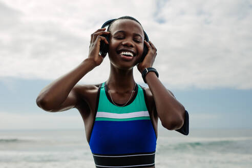 Lächelnde Frau mit Kopfhörern, die während einer Trainingspause Musik hört. Frau genießt das Hören von Musik nach dem Training im Freien. - JLPSF14810
