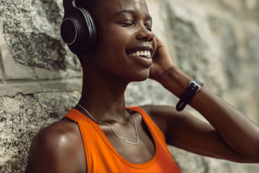 Close-up of a woman enjoying listening music while having break outdoors after training session. Female athlete with headphones relaxing by a wall after workout - JLPSF14815