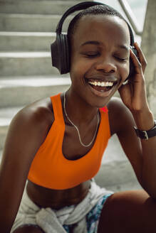 Smiling woman sitting on steps with headphones resting after workout. Fit female athlete taking a break after exercising enjoying listening music. - JLPSF14816