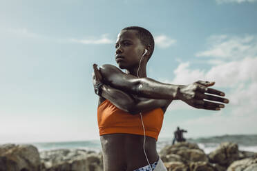 Afrikanerin mit Kopfhörern, die ihre Arme im Freien ausstreckt. Frau in Sportkleidung beim Sport am Strand. - JLPSF14826