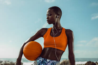 Afrikanische Frau in Sportkleidung, die einen Basketball hält, steht am Strand. Fitness-Frau mit Kopfhörern, die nach dem Sporttraining Musik hört. - JLPSF14828