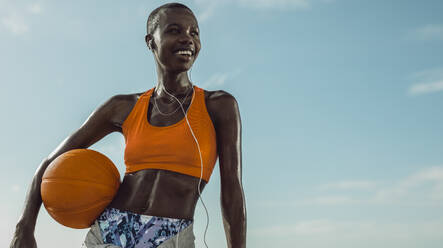 Frau in Fitnesskleidung, die im Freien steht und einen Basketball hält. Basketballspielerin, die wegschaut und gegen den Himmel lächelt. - JLPSF14829