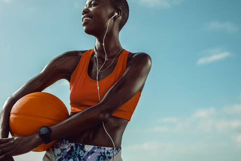 Sportlerin, die im Freien steht und einen Basketball hält. Frau mit Basketball, die wegschaut und gegen den Himmel lächelt. - JLPSF14830