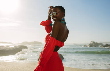 Schöne afrikanische Frau in rotem Kleid genießt am Strand. Lächelnde Frau in schönem Kleid spaziert am Ufer entlang. - JLPSF14831