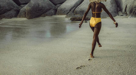 Rear view of a african woman wearing yellow bikini walking along the seashore. Woman walking on the beach in swimwear. - JLPSF14835