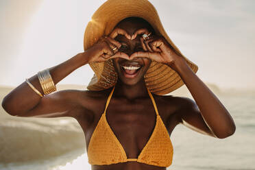 Fröhliche Frau im Bikini, die am Strand mit ihren Fingern ein Herz macht. Afrikanische Frau mit Strohhut, die am Meer ein Herz mit den Händen macht. - JLPSF14842