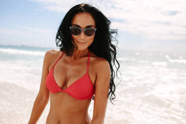 Lächelnde Frau am Strand. Frau im Bikini mit Sonnenbrille am Strand stehend mit Meer im Hintergrund. - JLPSF14855