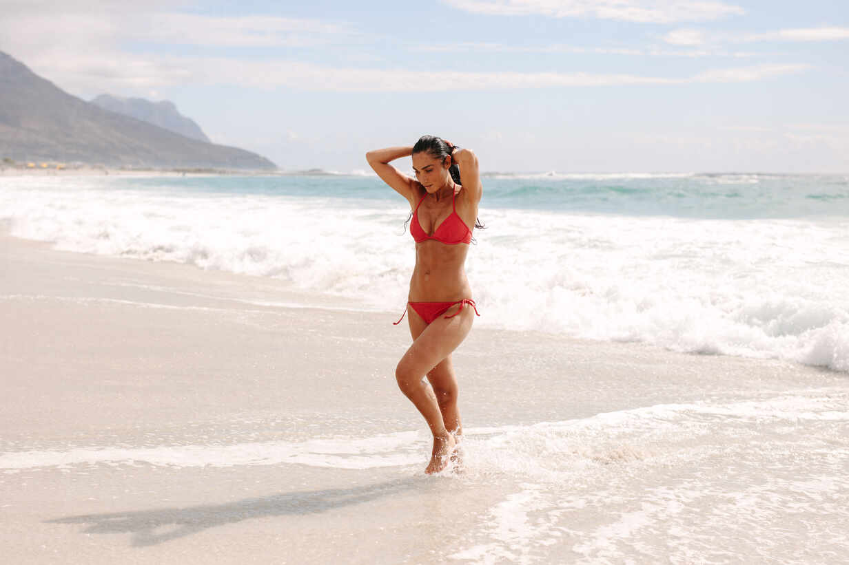 Beautiful smiling woman wearing red thongs and black bikini posing on the  sea beach at bright sunny day against ocean waves. Girl relaxing and having  good time during summer holiday vacation. Stock